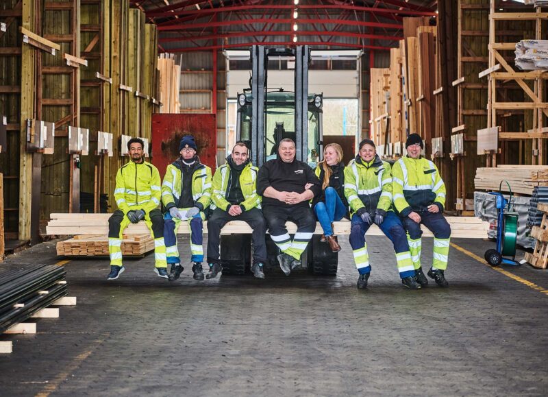 Vi Bygger Vejle: Et team på syv medarbejdere fra trælasten smiler, siddende på træ i lagerhal med gaffeltruck.