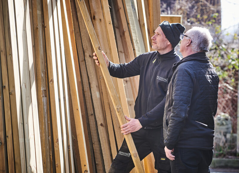 Vi Bygger Vejle: To mænd inspicerer træplanker. Den yngre holder en planke, den ældre ser på.