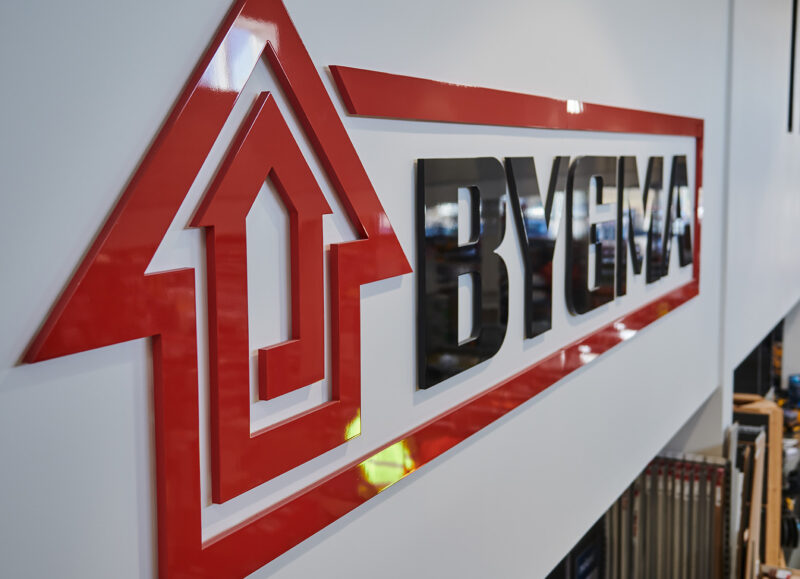 Vi Bygger Vejle: Bygma logo med rød husform og sorte bogstaver på hvid væg.