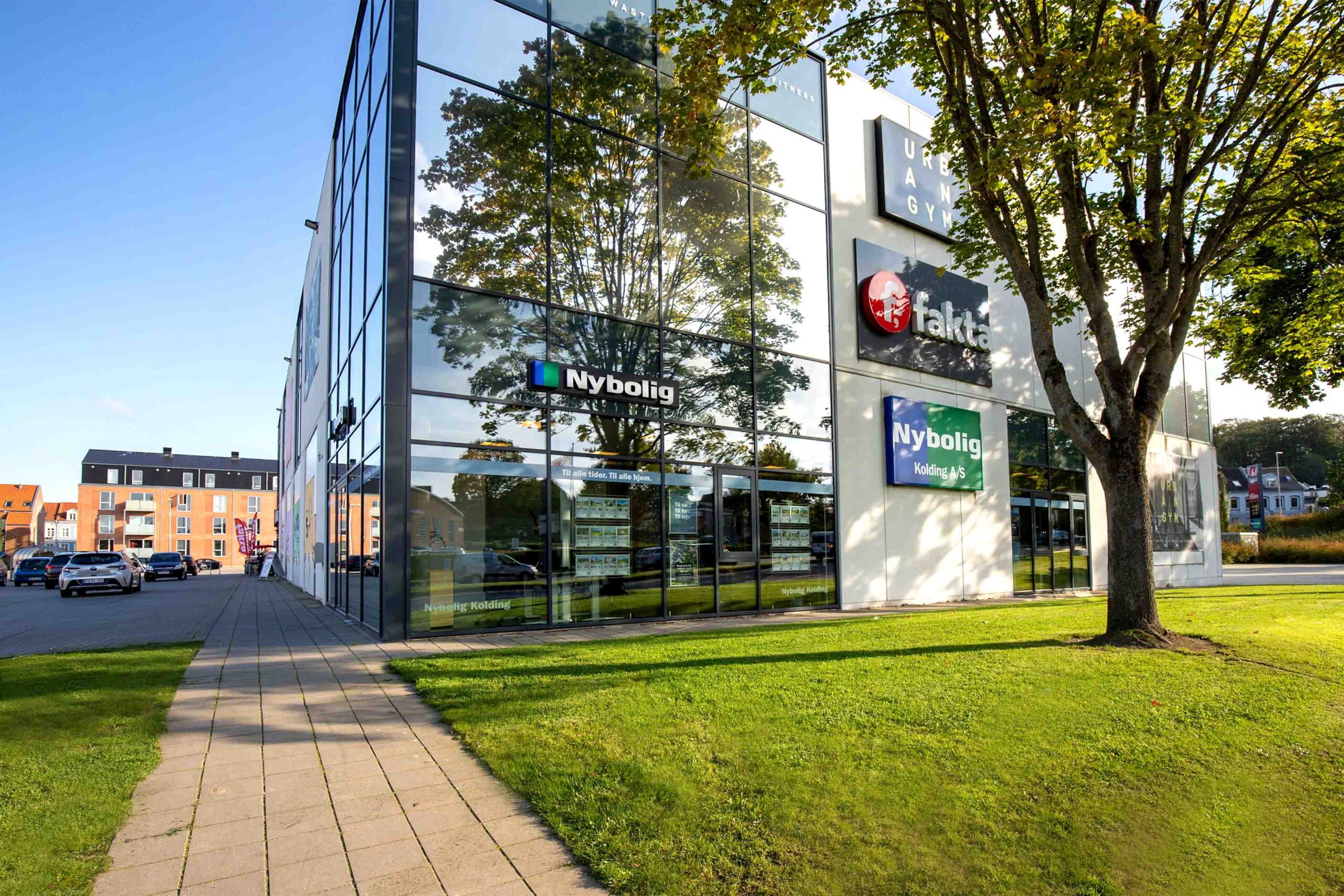 Vi Bygger Vejle: Moderne bygning med glasfacade, Nybolig, Fakta og Urban Gym, samt grønne omgivelser.