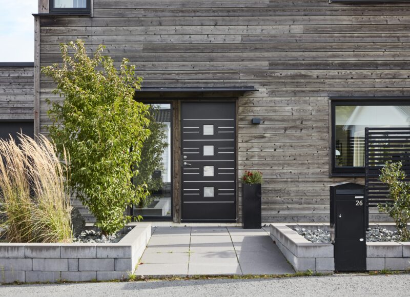 Vi Bygger Vejle: Moderne husfacade i træ, sort dør, frodige planter og sort postkasse nr. 26.