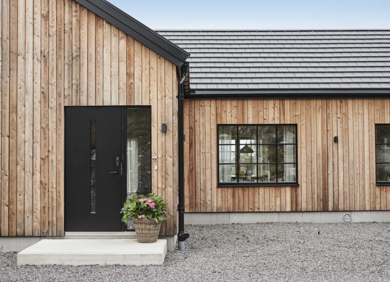 Vi Bygger Vejle: Moderne hus med træbeklædning, sort dør, sorte vinduer, gråt tag og blomsterkurv.