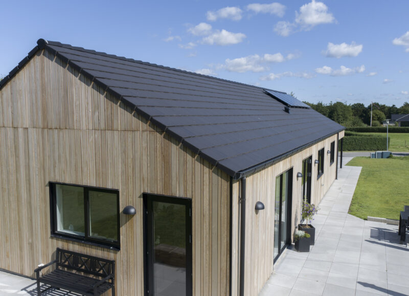 Vi Bygger Vejle: Moderne villa med træbeklædning, sort tegltag, solpaneler og en stor fliseterrasse.