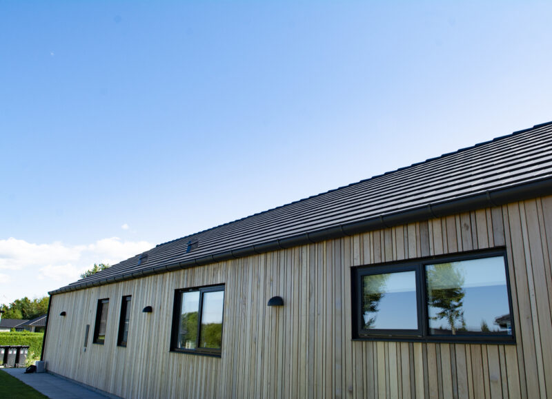 Vi Bygger Vejle: Moderne bygning med træbeklædning og mørkt tegltag under en klar blå himmel.