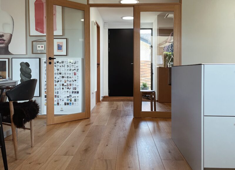 Vi Bygger Vejle: Entré med lyst plankegulv, glasdøre, kunst på væggen og en sort dør i gangen.