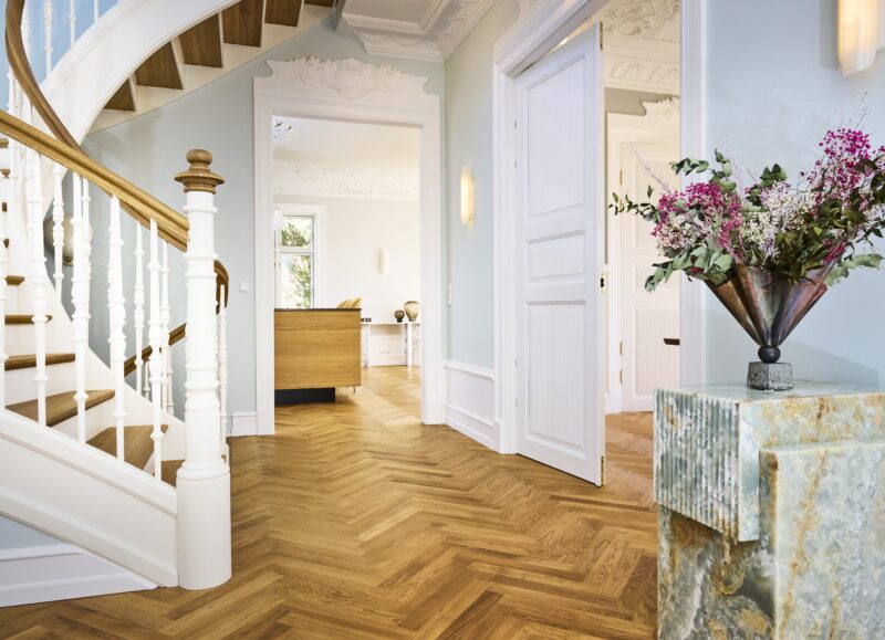 Vi Bygger Vejle: Elegant entré med buet trappe, sildebensparket og blomsterdekoration på marmorsokkel.