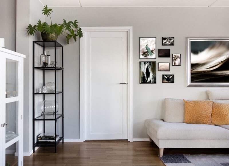 Vi Bygger Vejle: Moderne stue med grå vægge, hvid dør, sort reol, sofa med orange puder og billedvæg.