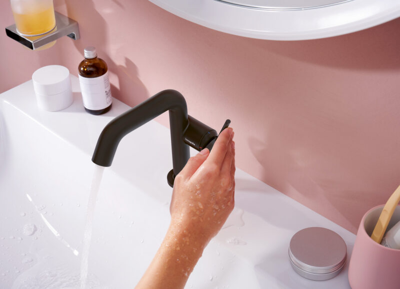 Vi Bygger Vejle: En våd hånd tænder for en sort Hansgrohe vandhane i en hvid vask med vand, der løber.
