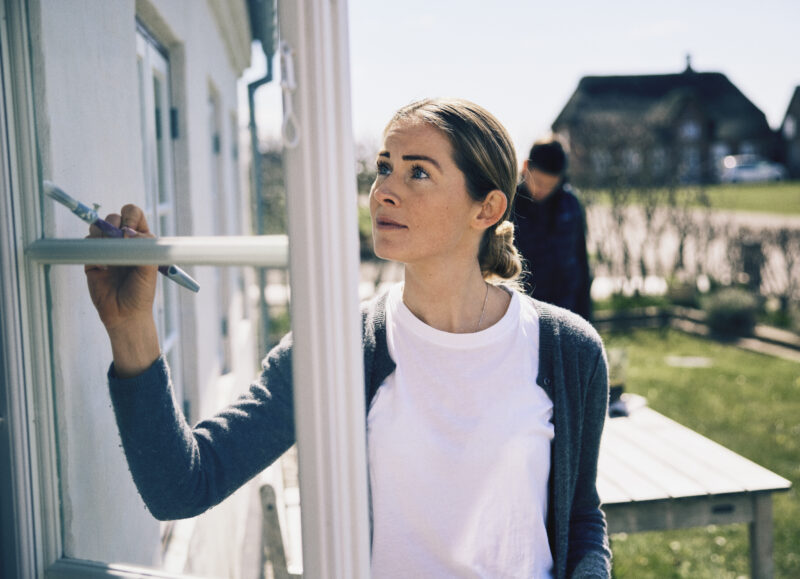 Vi Bygger Vejle: Kvinde maler vinduesramme på hus med stråtag i solskin.
