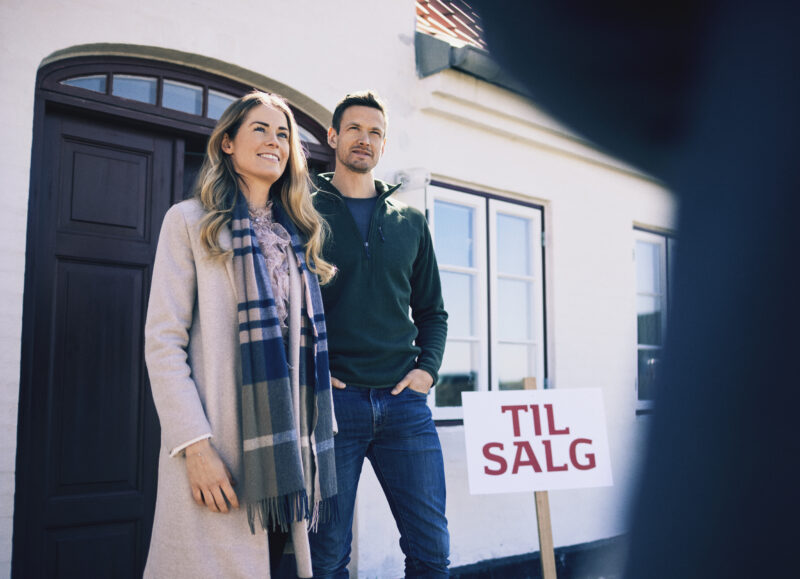 Vi Bygger Vejle: Smilende par på husjagt foran et hvidt hus med "Til Salg" skilt.