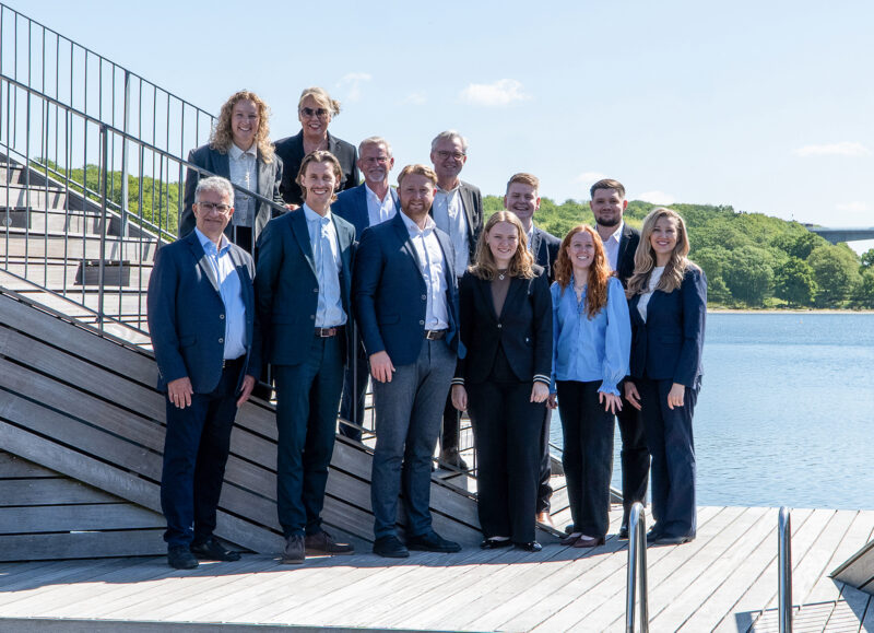 Vi Bygger Vejle: Teamet poserer smilende på en træterrasse ved vandet med Vejle Fjord Broen i baggrunden.