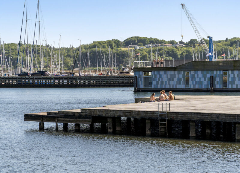 Vi Bygger Vejle: Mennesker solbader på en badebro med udsigt til en marina, flydende bygning og en kran.