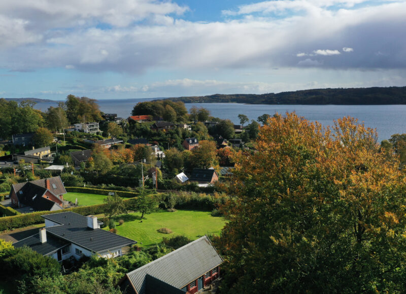 Vi Bygger Vejle: Efterårsbillede af parcelhuse ved Vejle Fjord, omgivet af farverige træer under en skyet himmel.
