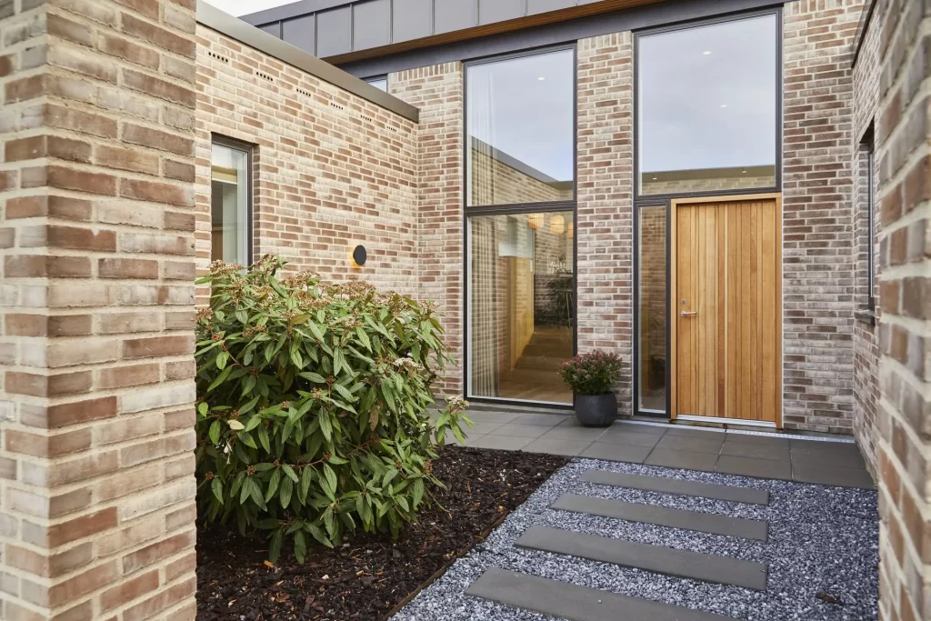 Vi Bygger Vejle: Moderne villa med lys murstensfacade, stor trædør, vinduer og sti af stenfliser og grus.