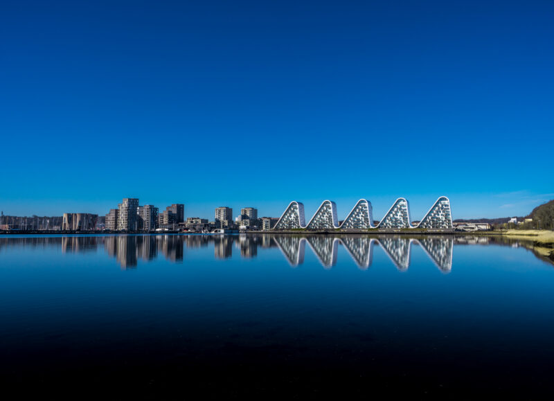 Vi Bygger Vejle: Bølgen og moderne lejligheder ved Vejle Fjord reflekteres i det stille vand under blå himmel.