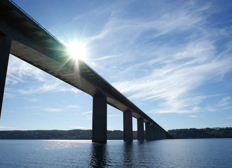 Vi Bygger Vejle: Solen stråler bag Vejlefjordbroen, set fra fjorden med træer i horisonten.