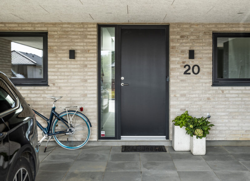 Vi Bygger Vejle: Moderne indgangsparti med lyse mursten, sort dør, nummer 20, cykel, bil og grønne planter.