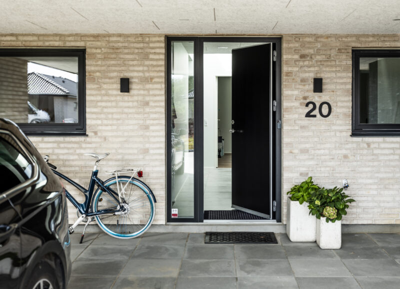 Vi Bygger Vejle: Moderne entré med åben sort dør, nr. 20, lys mursten, en cykel og bil.