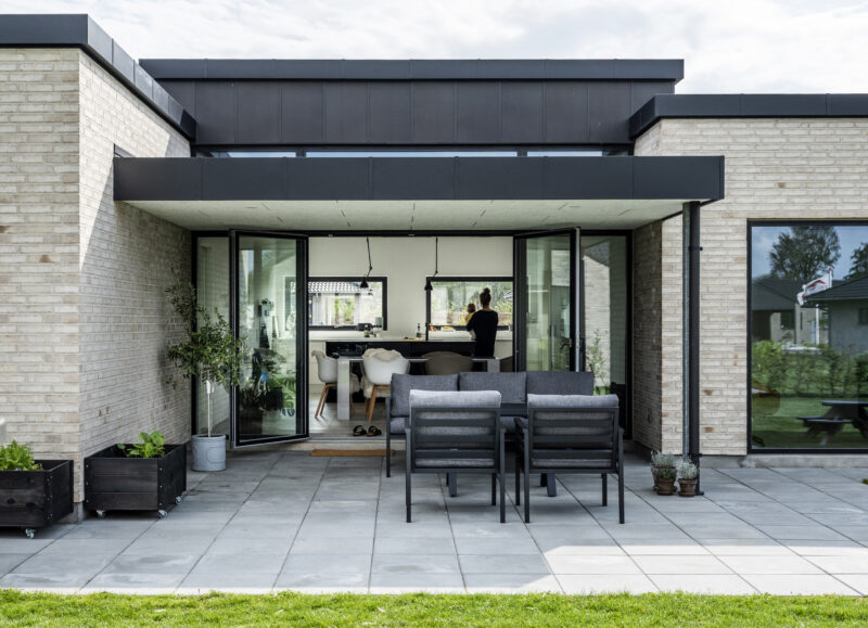 Vi Bygger Vejle: Moderne parcelhus med lys mursten, sort tag, stor terrasse og åbent køkken-alrum.