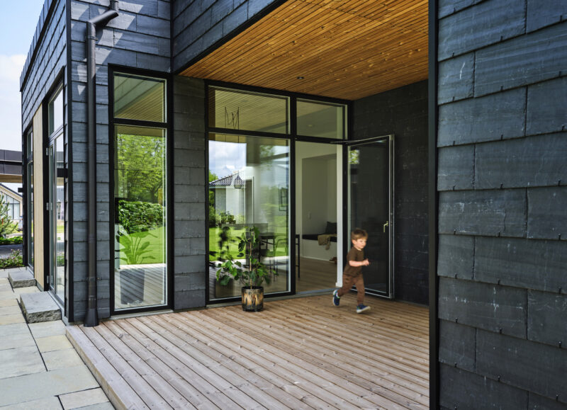 Vi Bygger Vejle: Moderne hus med sort skiferfacade og træterrasse, hvor et barn løber ud.