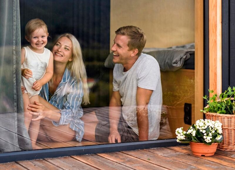 Vi Bygger Vejle: Smilende familie med lille barn i moderne hus' store vindue. Træterrasse udenfor.