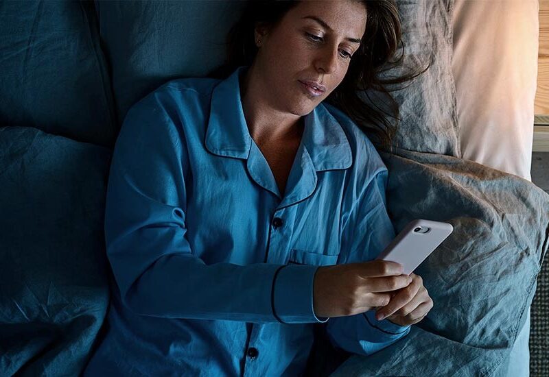 Vi Bygger Vejle: En kvinde i blå pyjamas ligger i sengen og ser på sin telefon i mørket med en lampe tændt.