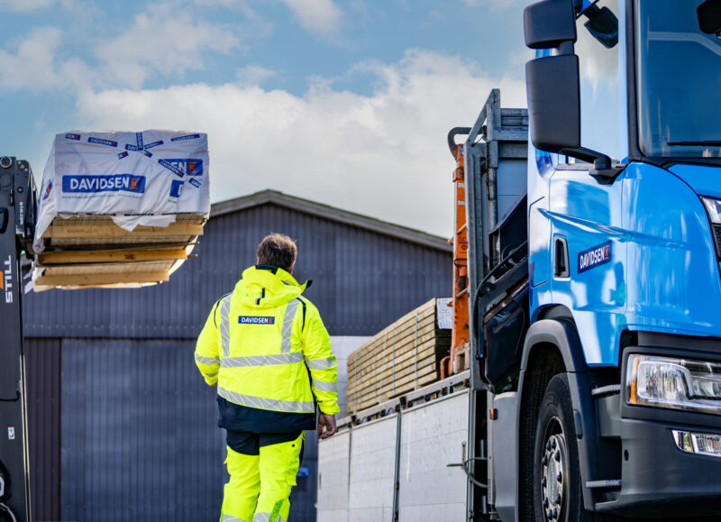 Vi Bygger Vejle: En arbejder i gul jakke går ved en Davidsen lastbil, mens en STILL gaffeltruck løfter materialer.