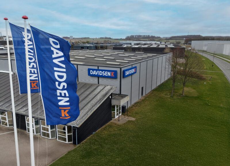 Vi Bygger Vejle: Luftfoto af Davidsen Vejle byggecenter med blå flag og bygninger i industriområde.
