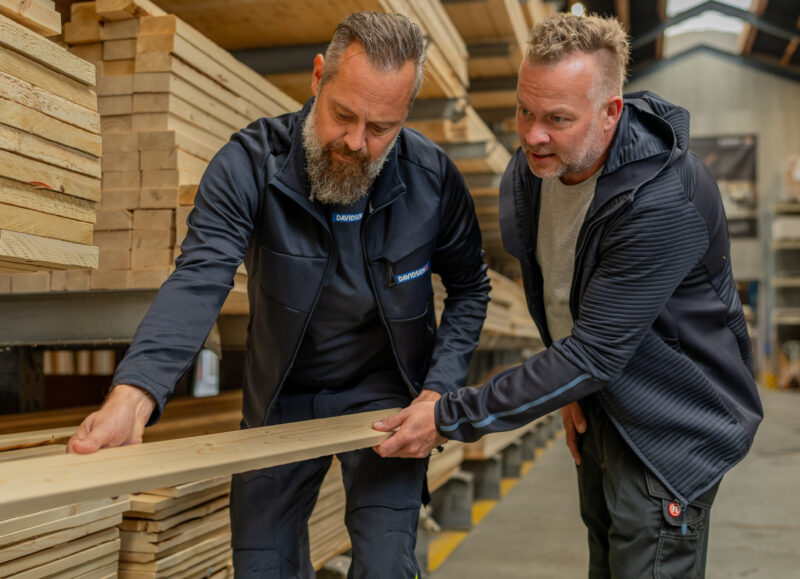 Vi Bygger Vejle: To mænd, den ene med Davidsen-logo, kigger på en træplanke i et lager.