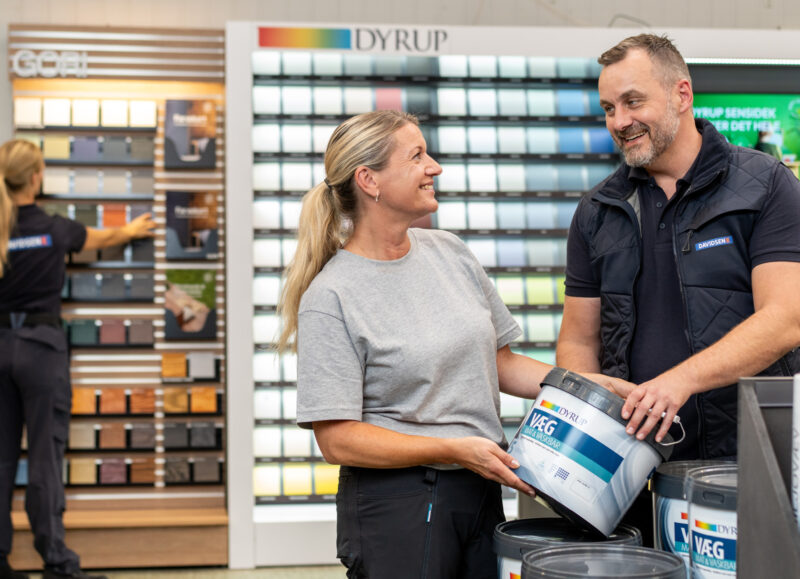 Vi Bygger Vejle: En mand og en kvinde smiler og taler, mens de holder en malerbøtte i en farvehandel. Farveprøver i baggrunden.