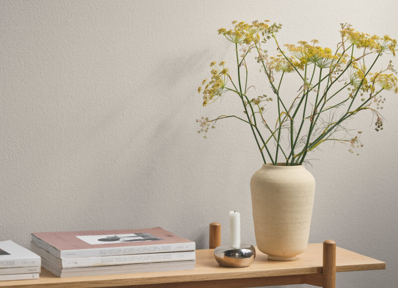 Vi Bygger Vejle: Indretningsdetalje med lys væg, træhylde, bøger, vase med gule dildblomster og lysestage med hvidt lys.
