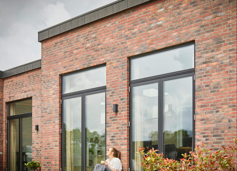 Vi Bygger Vejle: Moderne murstenshus med store vinduer og en kvinde der sidder på terrassen med en kop.