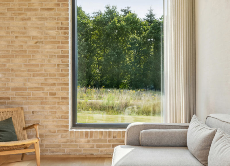 Vi Bygger Vejle: Moderne stue med lys murstensvæg, stor vinduesudsigt til grøn natur, træstol og lys sofa.