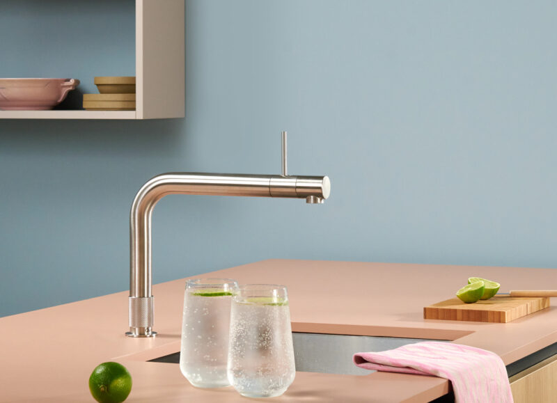 Vi Bygger Vejle: Moderne køkken med rosa bordplade, stålhane, to glas vand med lime, lyserød karklud og lyseblå væg.