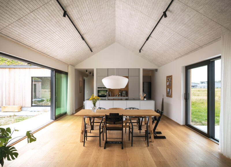 Vi Bygger Vejle: Lyst, moderne køkken-alrum med trægulv, akustikloft, spisebord og store glasdøre til terrasse og have.