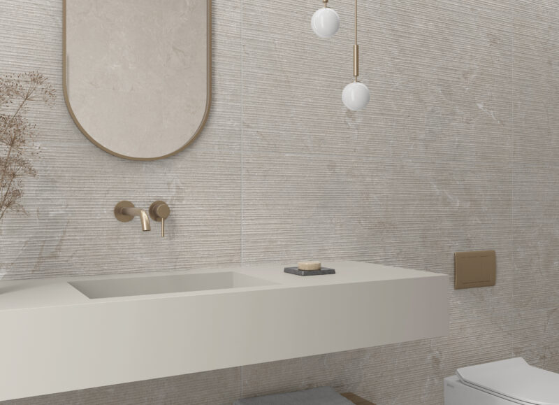 Vi Bygger Vejle: Elegant badeværelse med strukturerede beige fliser, flydende håndvask, messingarmatur, ovalt spejl og toilet.